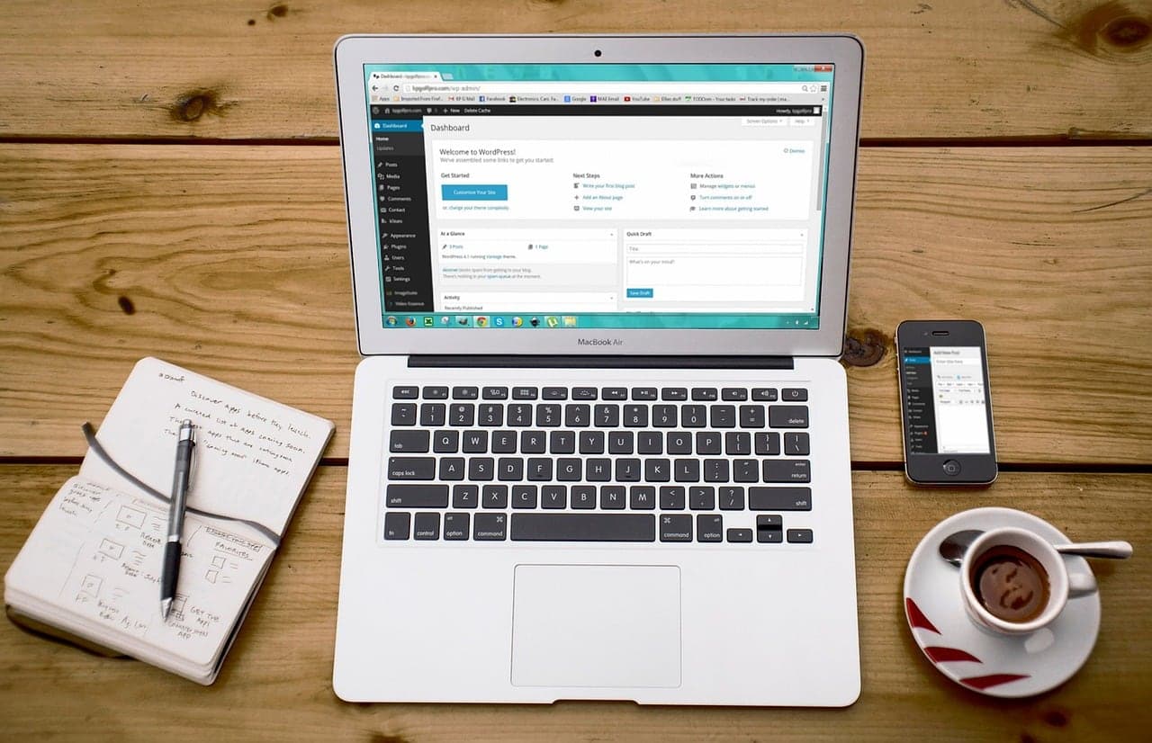 A laptop showing a website on a wooden table with a notebook and coffee
