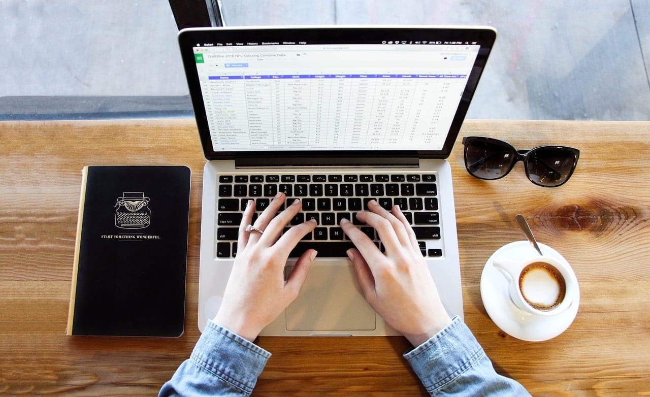 Hands typing on a laptop showing a spreadsheet beside a coffee